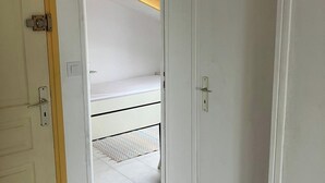 Une porte blanche donne sur une pièce qui contient un lit, une petite fenêtre et un plafond avec une poutre en bois.