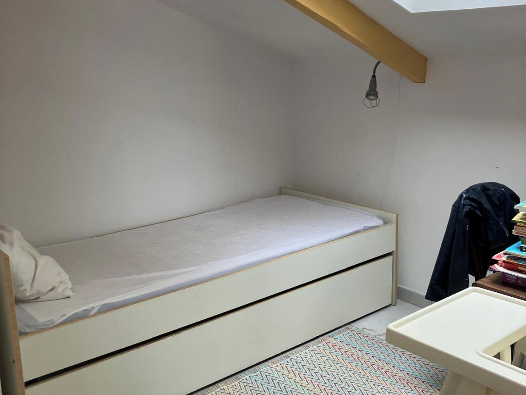 A small, single-bed room with a simple design, a desk with a chair, and a small table with books.