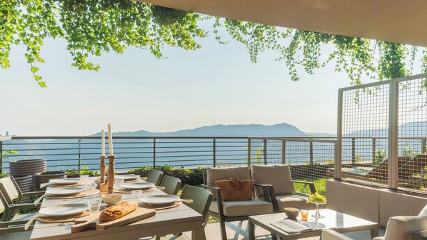 An outdoor dining area with tables set for a meal, overlooking a body of water and hills. - Skyline Lake Apartment (Como)