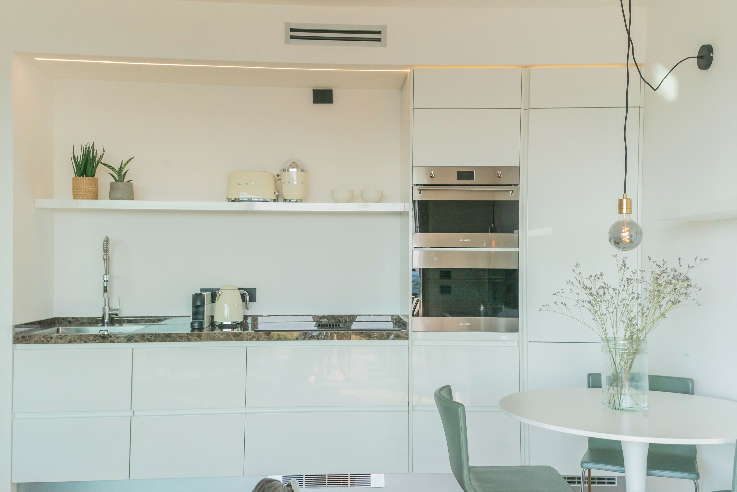 A modern kitchen with white cabinets, a built-in oven, and a round dining table with chairs.