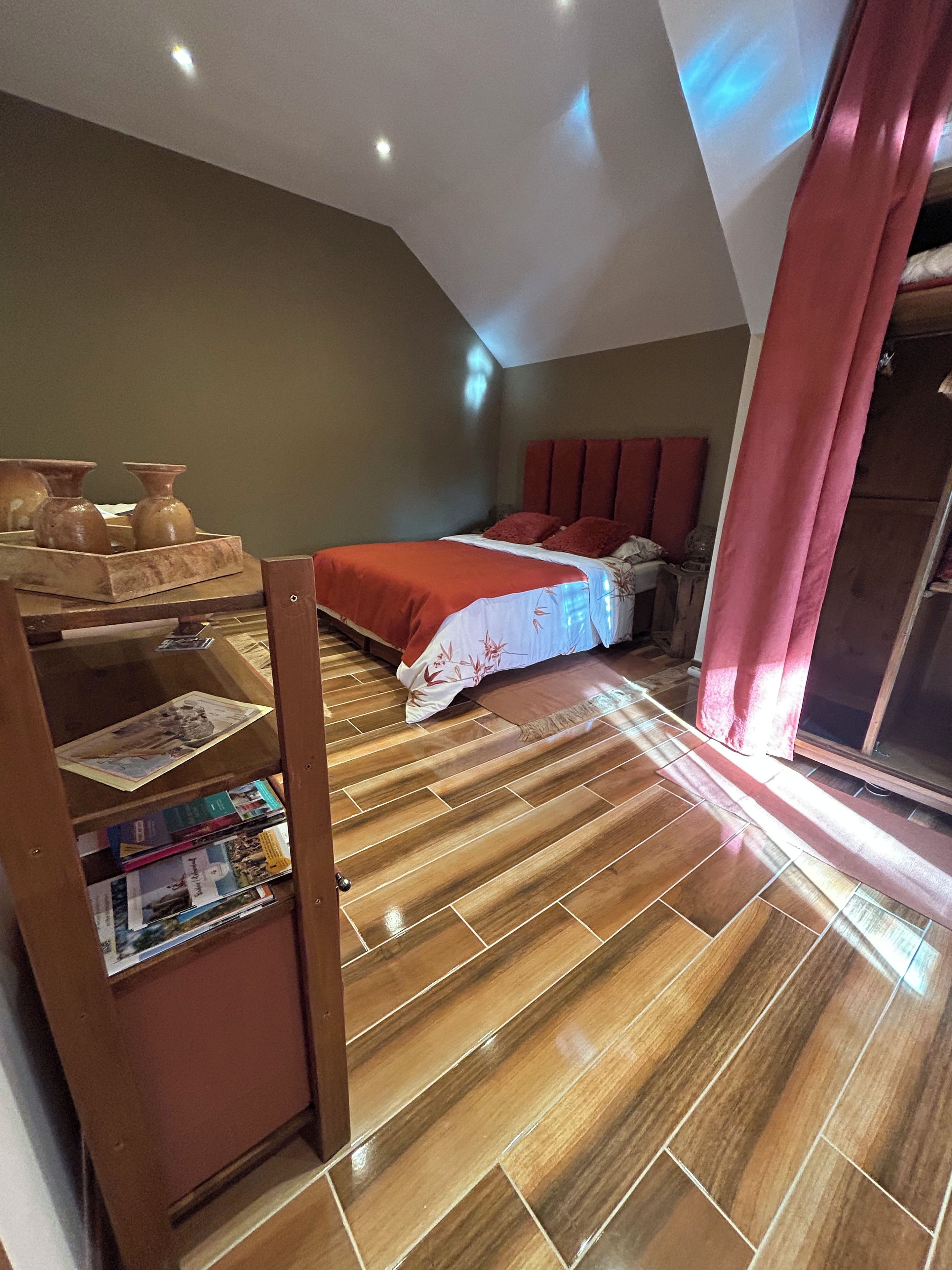 A bedroom with a wooden floor, a bed with a red bedspread, and a shelf with books.