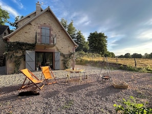 A rustic stone house with a balcony, outdoor seating, and a gravel ground.