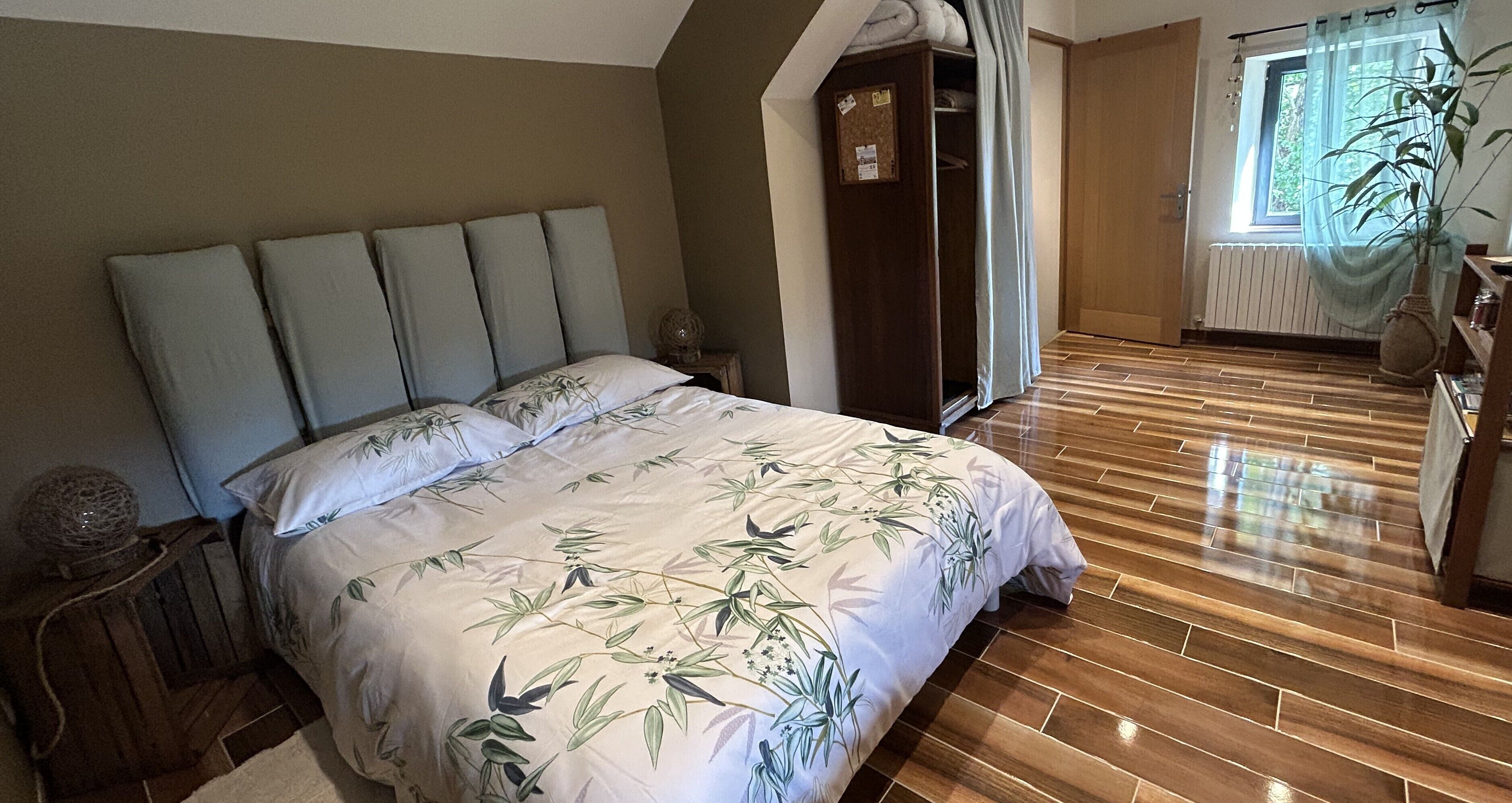 Un dormitorio con una cama, suelo de madera, un armario, una ventana con una planta y una puerta.