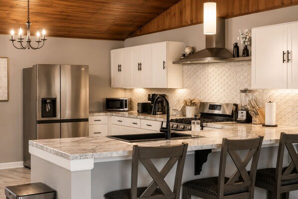 A modern kitchen with stainless steel appliances, white cabinetry, a marble island, and a wooden ceiling.