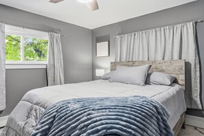 A bedroom with a bed, a window with curtains, a ceiling fan, and a bedside table.