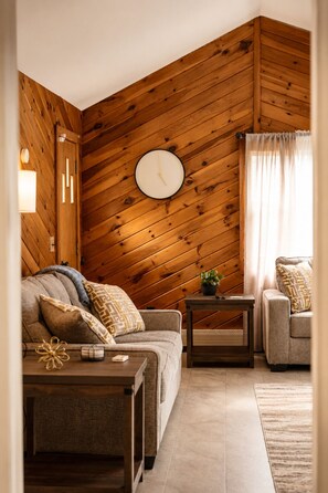 A cozy living room with a wooden sloped ceiling, a round wall clock, a sofa, a coffee table, and a side table with a plant.