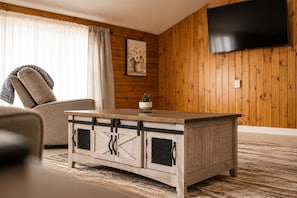 Un salon avec une table basse en bois, un canapé, un téléviseur fixé au mur et une fenêtre avec des rideaux.