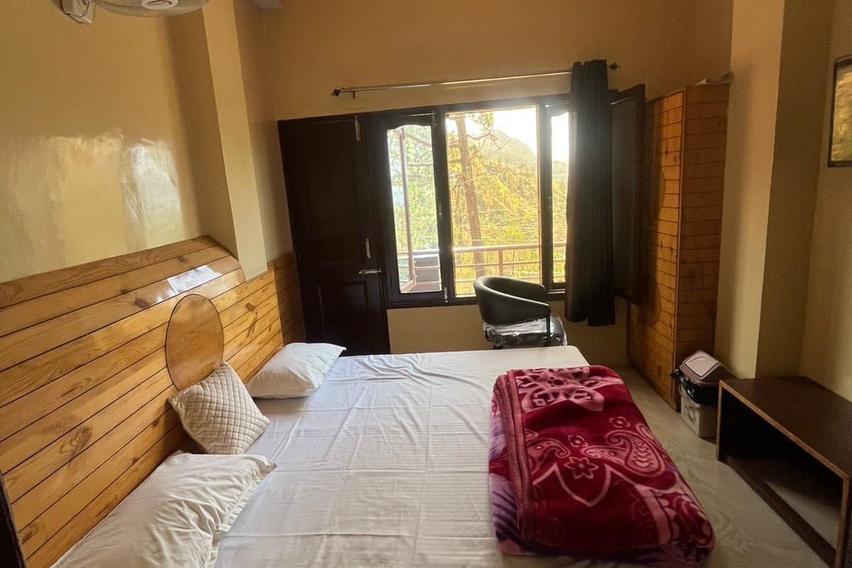 A bedroom with a wooden headboard, a bed with white linens, a red blanket, a ceiling fan, a window with a view of trees, and a small table.