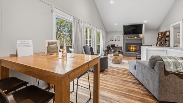 A modern living room with a wooden dining table, a grey sofa, a fireplace, and a television. - Forest Chalet With Hot Tub Near Gunstock 3 Bedroom Home (Gilford)