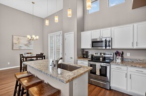 A kitchen with white cabinets, a granite countertop, stainless steel appliances, and a dining area with wooden chairs and a table. - Scottsdale Basecamp With Prime Golf Access 2 Bedroom Condo (Scottsdale)