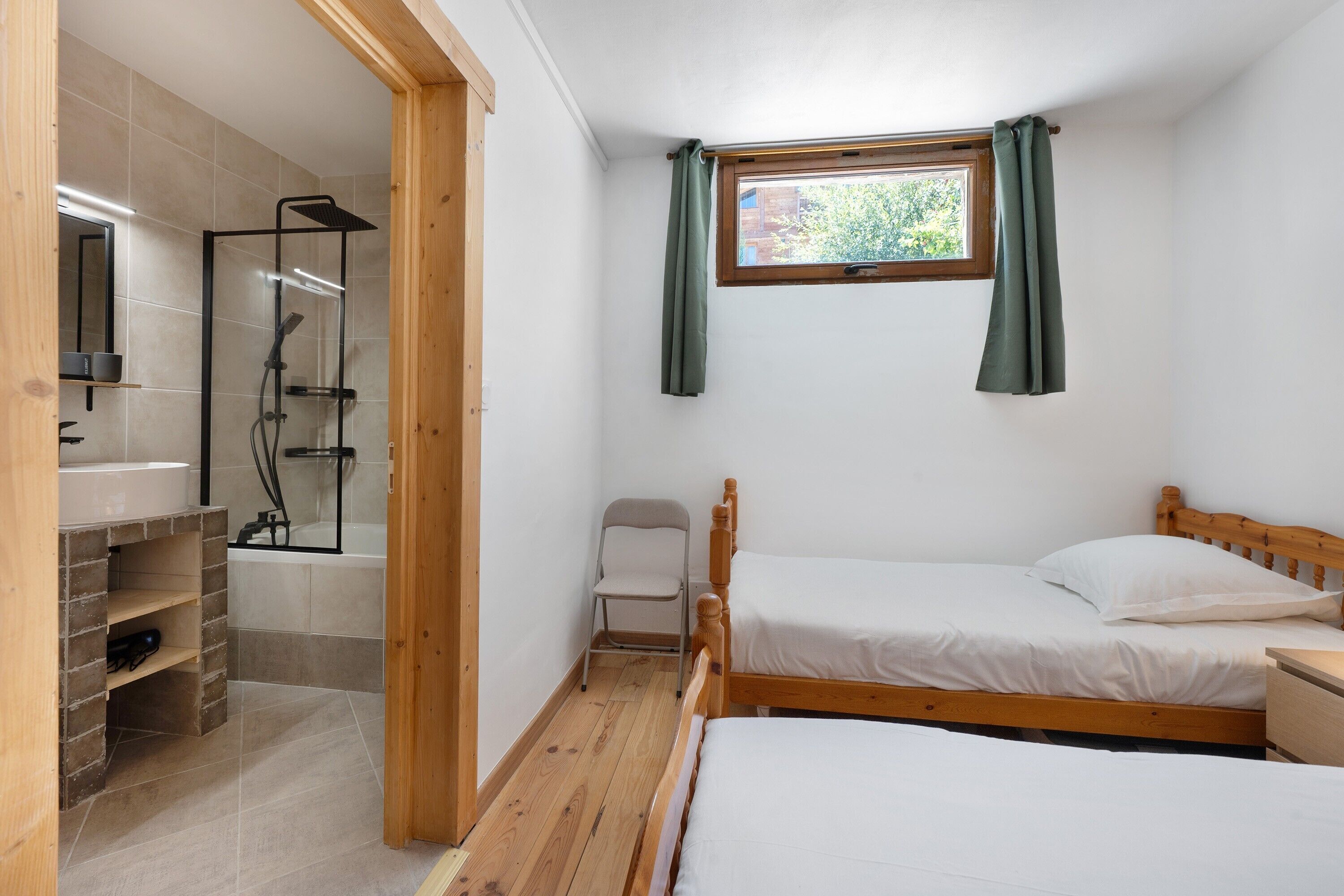 A bedroom with a wooden bed, a small window with curtains, a wooden chair, and a shower area.