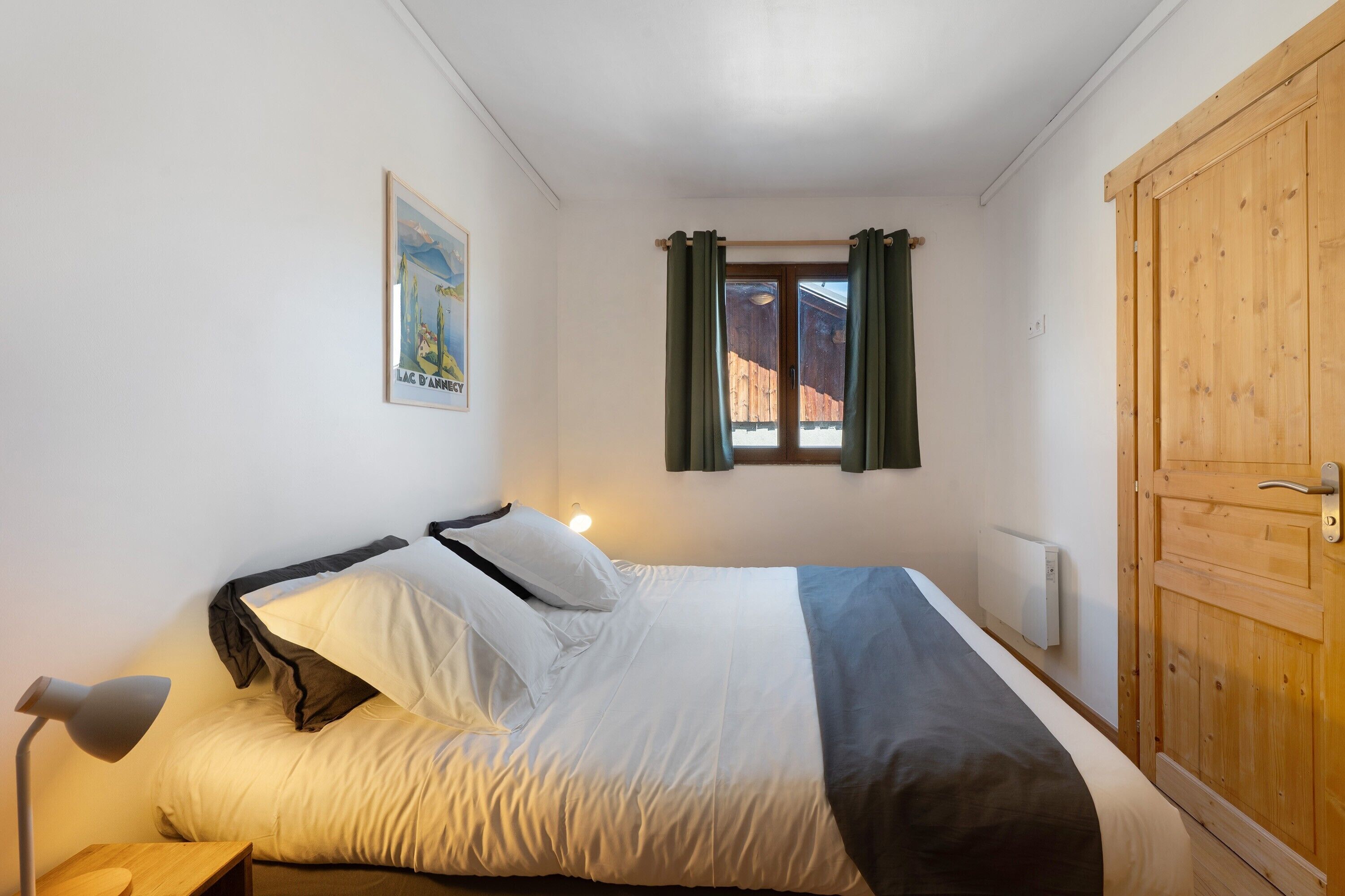 A bedroom with a bed, a nightstand, a lamp, a window with curtains, a wooden wardrobe, and a poster on the wall.