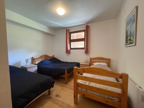 Una habitación pequeña y bien iluminada con dos camas de madera, una mesita de noche con lámpara, una ventana con cortinas y un cuadro colgado en la pared.