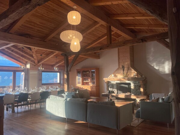 Una habitación espaciosa con techo alto, vigas de madera, chimenea de piedra, zona de comedor con mesas y sillas, y un rincón de estar acogedor con sofás.
