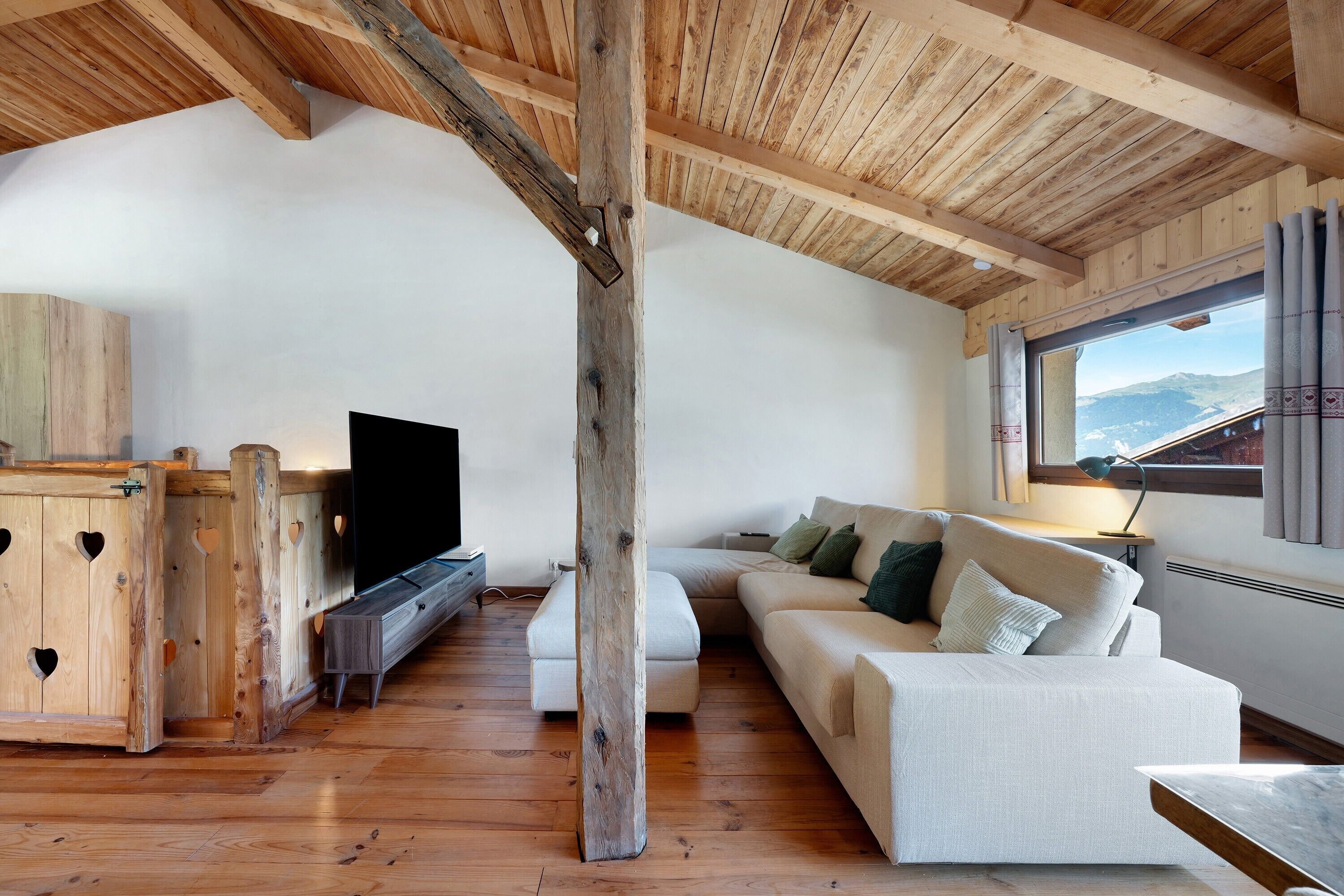 Ett rum i loftstil med ett träpanelat tak, en vit soffa, en tv och ett stort fönster med utsikt över bergen.
