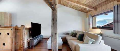A loft-style room with a wooden ceiling, a white sofa, a TV, and a large window with a mountain view.