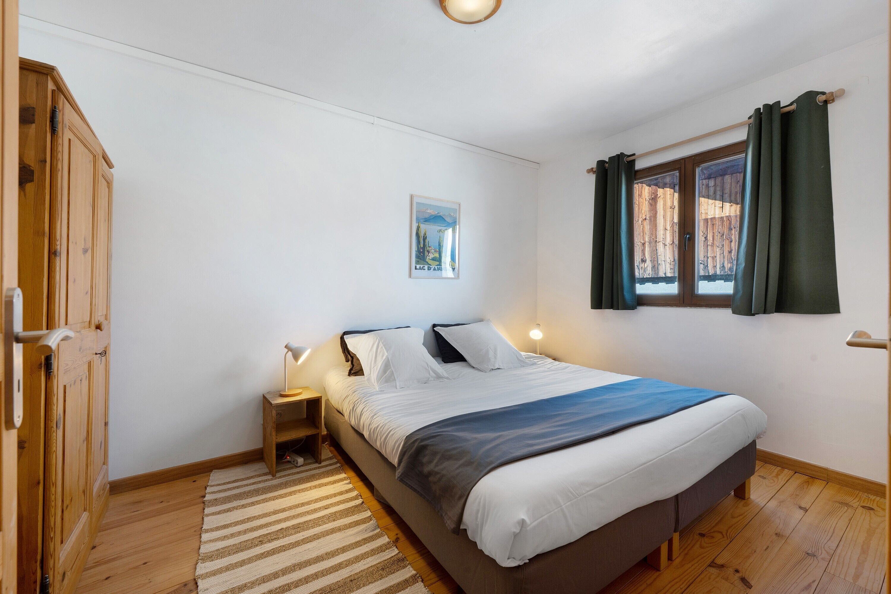 A bedroom with a bed, bedside tables, a wooden wardrobe, a window with curtains, and a wall-mounted picture.