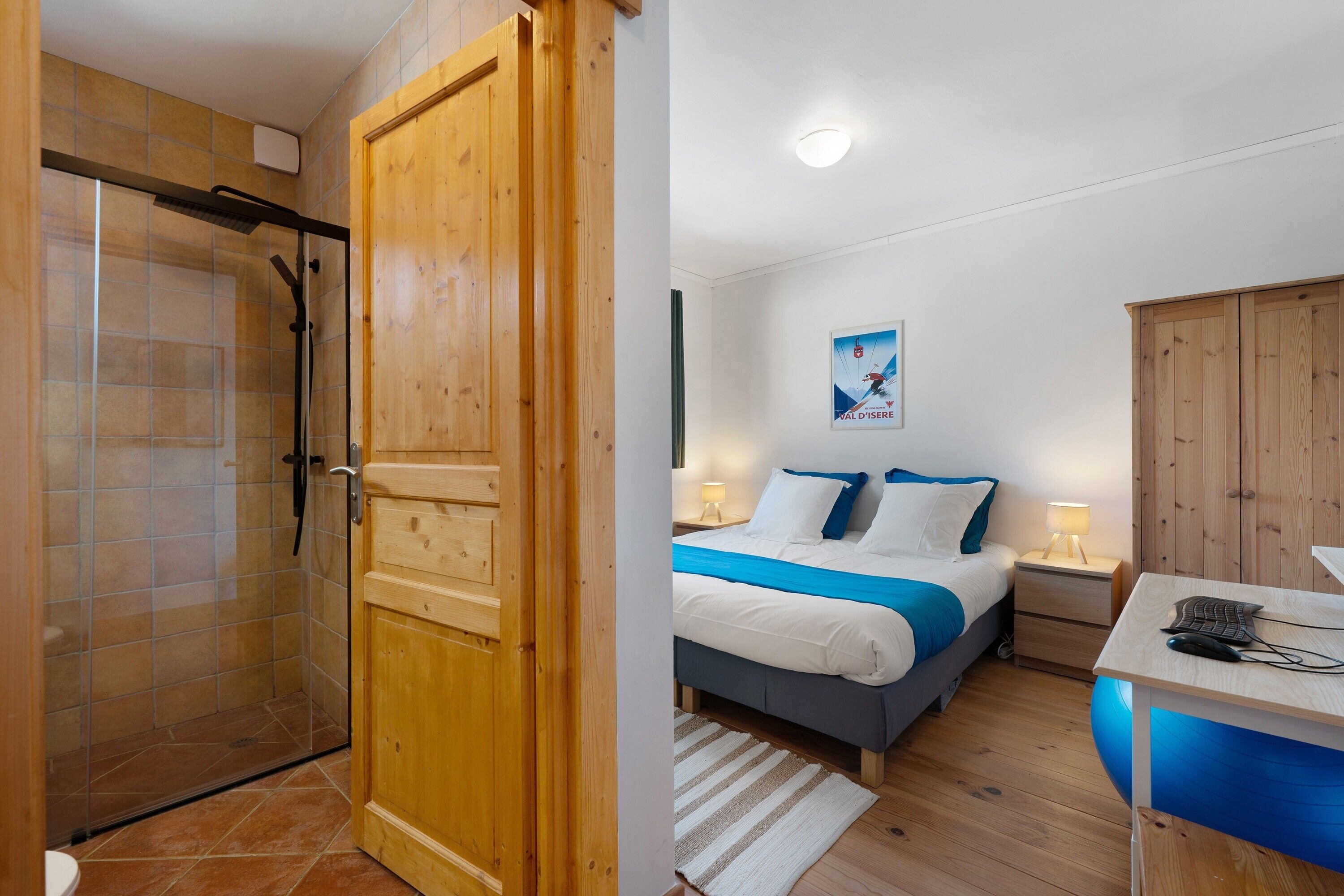 A hotel room with a shower, a bed with a blue bedspread, a desk with a computer, and a wooden wardrobe.