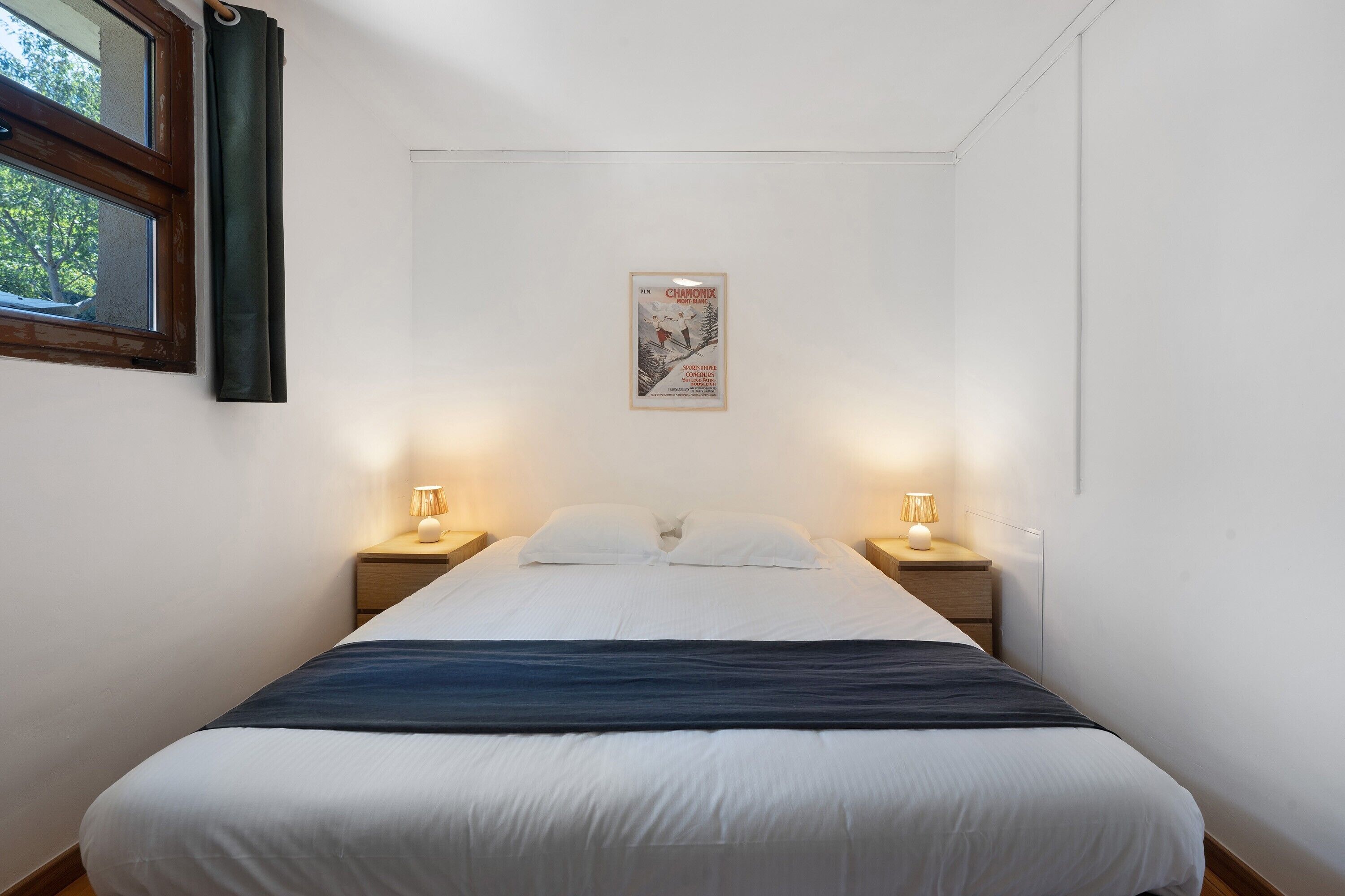 A bedroom with a bed, two bedside tables with lamps, a window with curtains, and a framed picture on the wall.