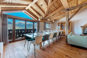 Ein Essbereich mit einem langen Tisch, gedeckt für eine Mahlzeit, große Fenster mit Blick auf die Berge, Holzbalken an der Decke und eine gemütliche Sitzecke mit einer Couch.
