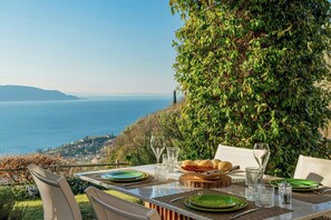 Ein Esstisch mit Meerblick, im Hintergrund ein Weinberg und ein üppiger, grüner Heckenstreifen.