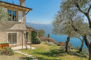 Ein Steinhaus mit Balkon, Terrasse und Blick auf einen See und Berge.