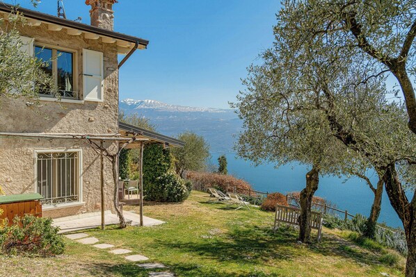 Ein Steinhaus mit Balkon, Terrasse und Blick auf einen See und Berge.