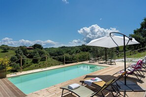 Ein Pool mit Liegestühlen und einem Sonnenschirm, umgeben von Grünflächen und blauem Himmel.
