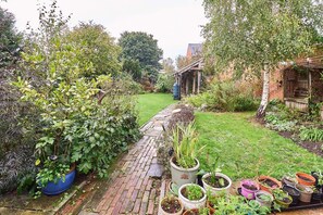 Ein Garten mit einem Ziegelweg, der zu einem rustikalen Gebäude führt, umgeben von verschiedenen Kübelpflanzen und Grünpflanzen.