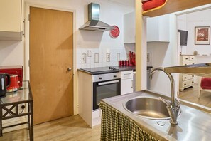 Eine kleine Küche mit Herd, Backofen und Spüle, ein roter Wasserkocher auf einem Tisch und eine Uhr an der Wand.