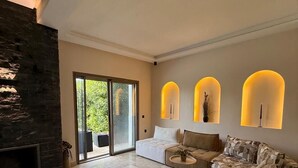 A spacious living room with a stone fireplace, a large sectional sofa, a coffee table, and a sliding glass door leading to an outdoor area. - Villa Silya – Private Pool Villa Steps from the Beach in Sidi Rahal (Sidi Rahal Chatai)
