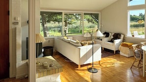Ein großzügiger Wohnbereich mit Holzboden, einem weißen Sofa, einem Couchtisch und einem großen Fenster mit Blick auf Grünflächen.