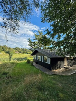 Eine kleine Holzhütte mit einer Terrasse, umgeben von üppigem Grün und wolkenlosem Himmel.