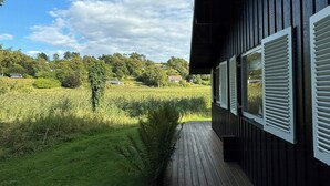 Eine Holzterrasse führt zu einem Gebäude mit Fensterläden, umgeben von einer Wiese und Bäumen.