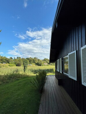 Eine Holzterrasse führt zu einem Gebäude mit Fensterläden, umgeben von einer Wiese und Bäumen.