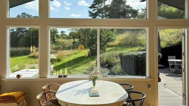 Ein Zimmer mit großem Fenster, einem runden Tisch, Korbstühlen und Blick auf eine grüne Landschaft.