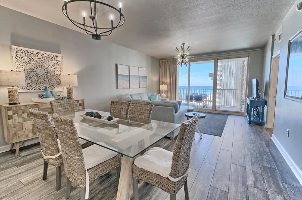 A modern living room with a glass dining table, wicker chairs, a grey sofa, and a balcony with a view.