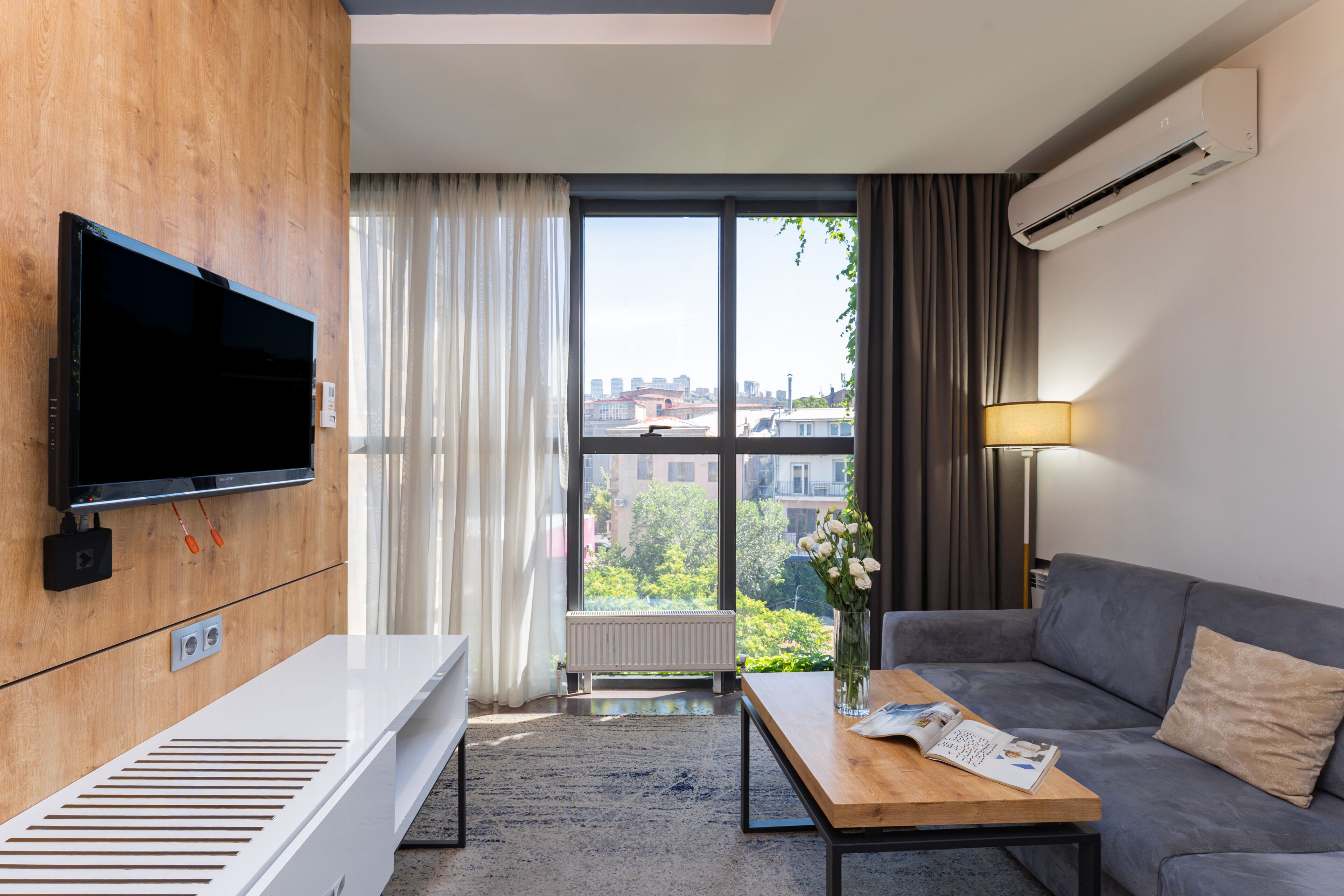 A modern hotel room with a flat-screen TV, a wooden panel wall, a sofa, a coffee table, and a view of the city through large windows.