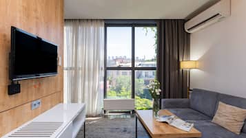 A modern hotel room with a flat-screen TV, a wooden panel wall, a sofa, a coffee table, and a view of the city through large windows.