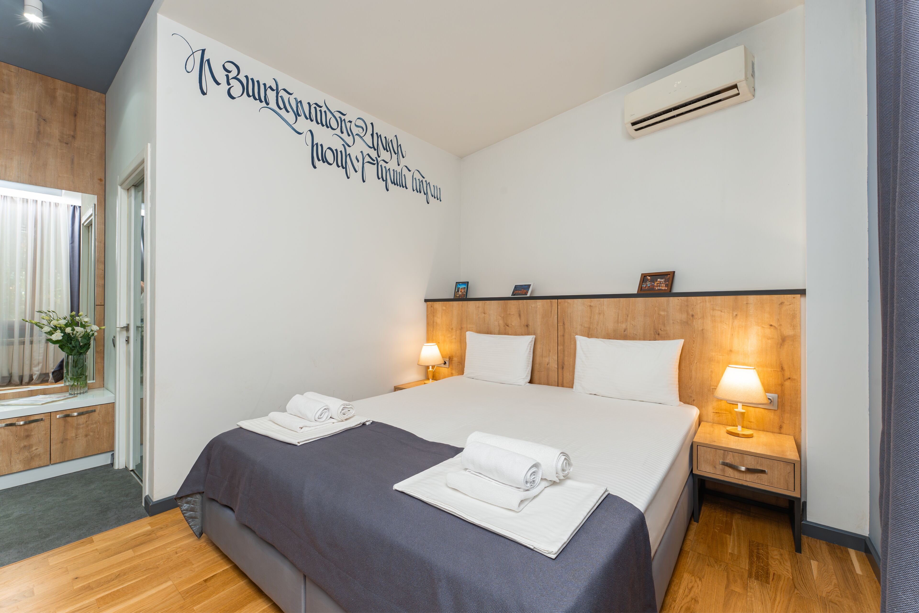 Quarto de hotel com uma cama grande, duas mesas de cabeceira com luminárias, uma cabeceira de madeira e uma parede com texto.