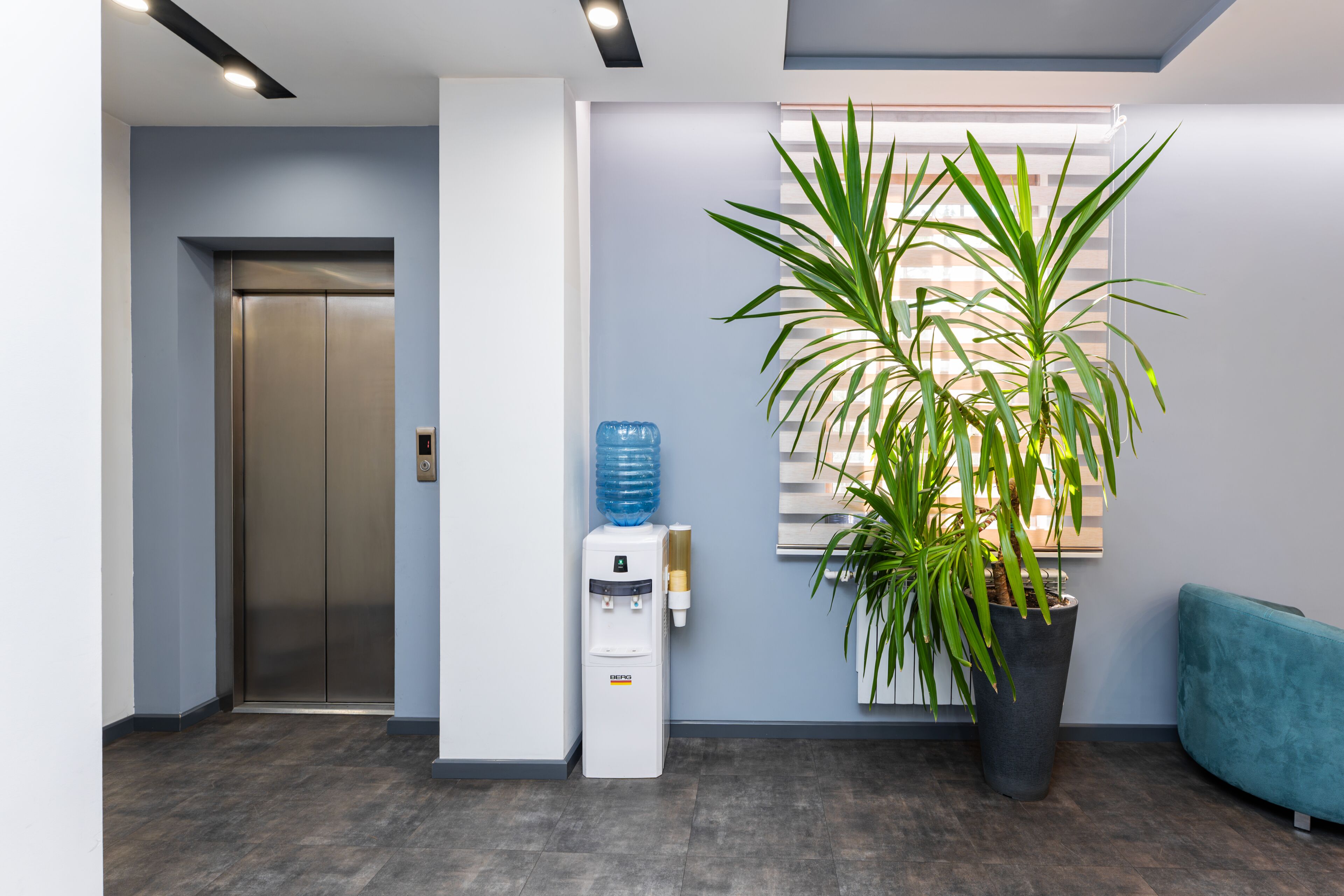 An elevator, water dispenser, potted plant, and teal seating area in a modern lobby.