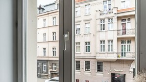 A window with a view of a street and buildings, a grey cushioned bench, and a door with a handle. - RentPlanet - Apartament Gliwicka (Katowice)