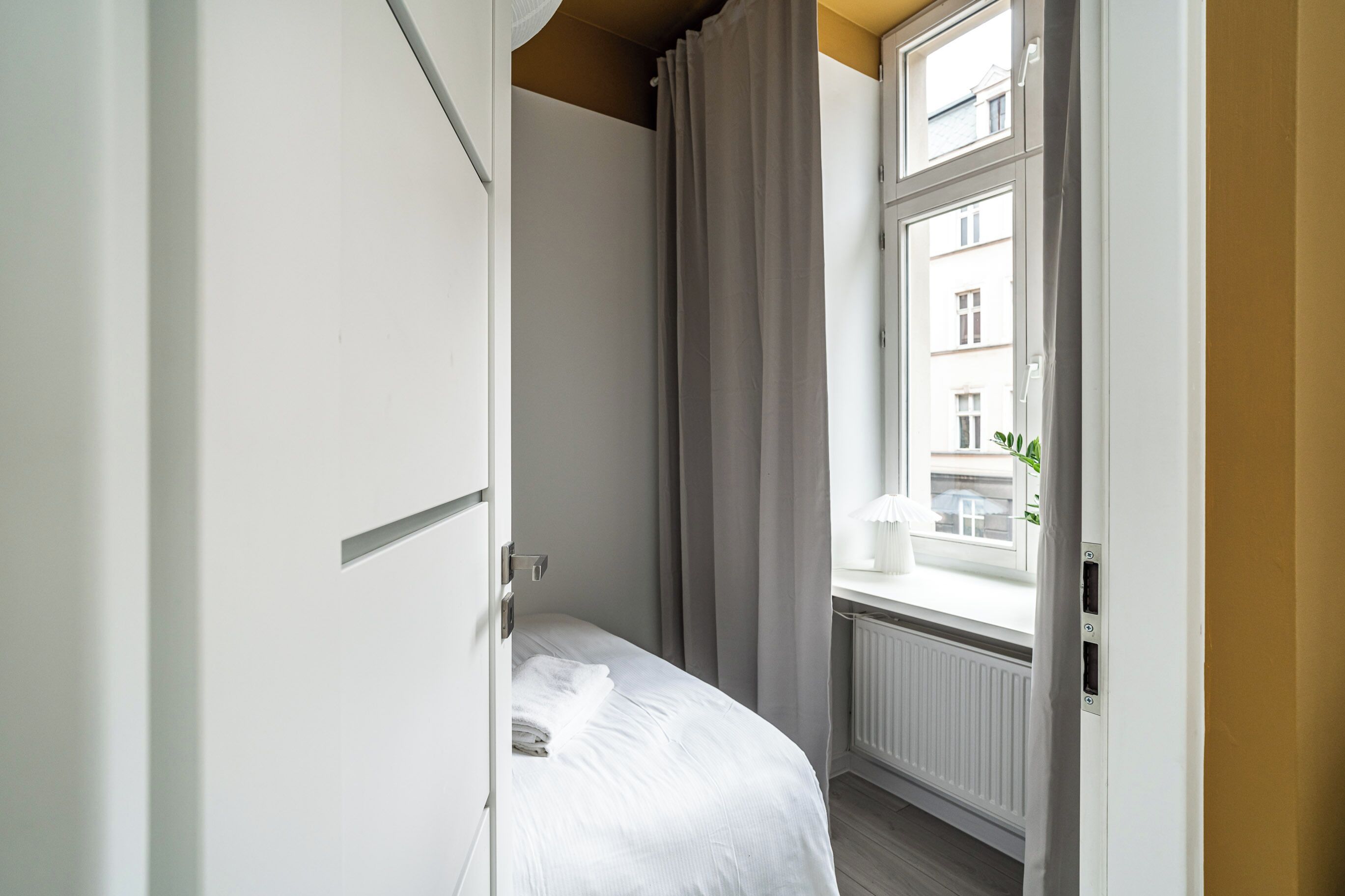 A hotel room with a bed, a window with curtains, a radiator, and a view of buildings outside.