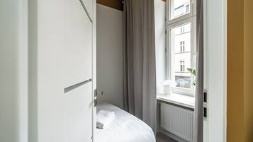 A hotel room with a bed, a window with curtains, a radiator, and a view of buildings outside.
