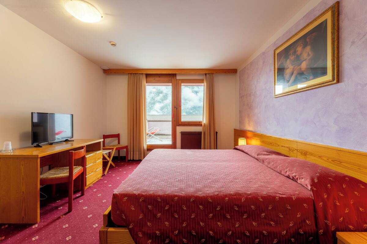 Una habitación de hotel con cama, escritorio, silla, televisor y ventana con cortinas.