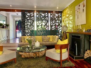 A living room with a sofa, chairs, a glass coffee table, a fireplace, and a decorative partition. - Our House, Your Home (Cape Town)