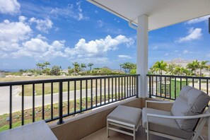 A balcony with a white chair and bench, a black railing, and a view of a road with palm trees and a distant mountain.