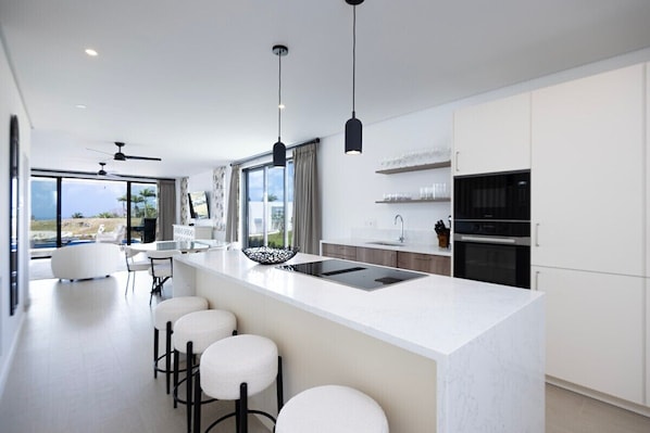 A modern kitchen with a central island, bar stools, a dining area with a view, and a large window.