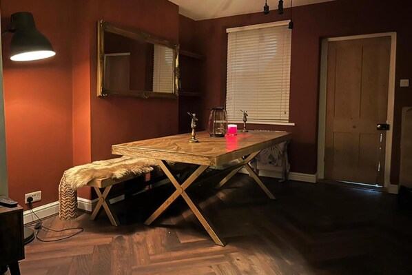 A room with a wooden table, a bench, a mirror, a window with blinds, and a door.