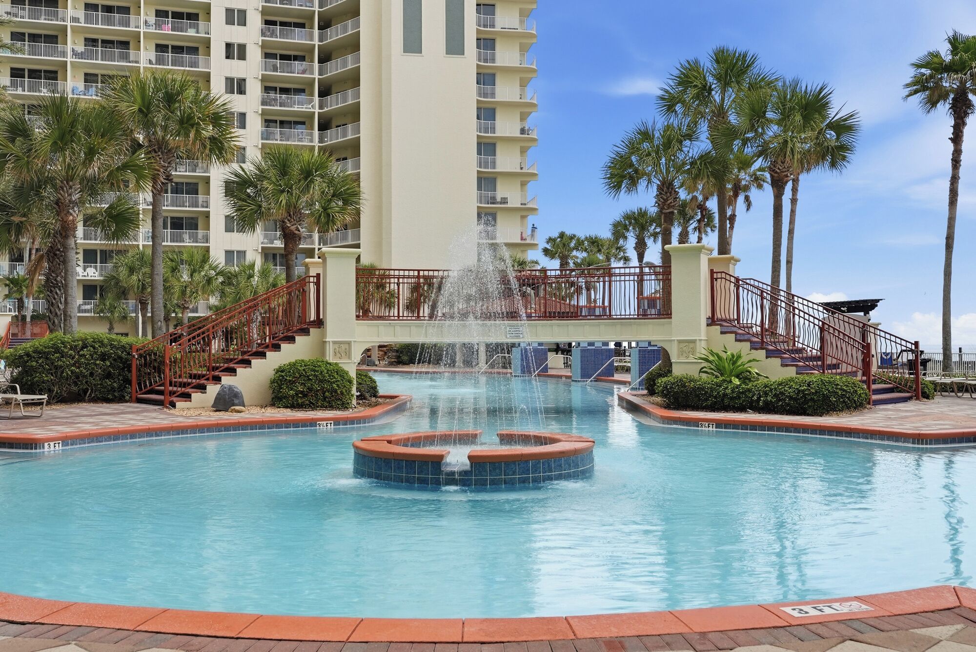 A hotel pool with a central water feature, surrounded by palm trees and a multi-story building with balconies.
